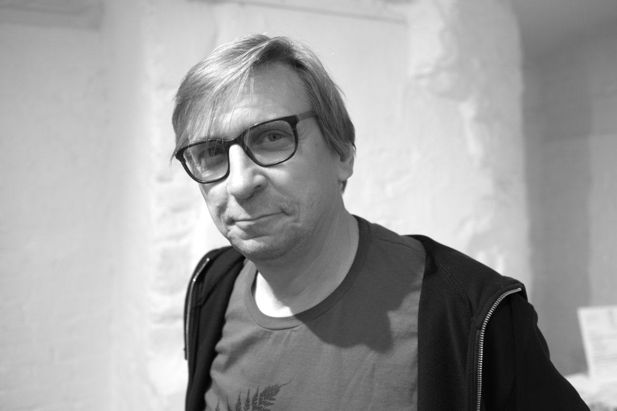 Schwarzweißes Porträtfoto im Querformat, das einen Mann mittleren Alters in T-Shirt und dunkler Jacke, mit dunkler Hornbrille zeigt, der leicht süffisant in die Kamera schaut.