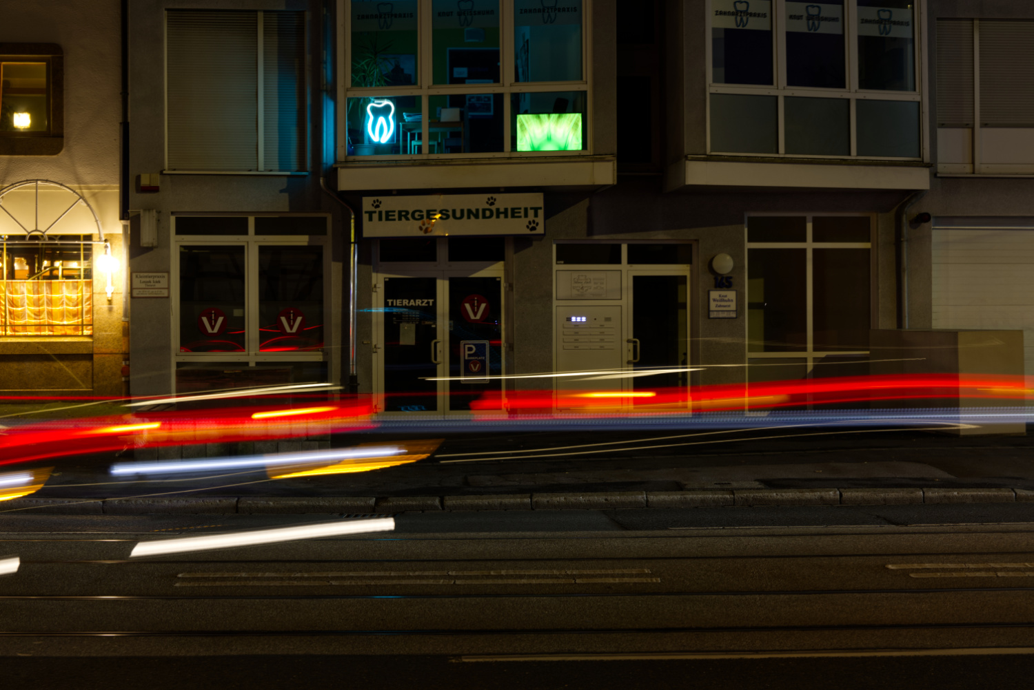 Querformatiges Farbfoto eines Gebäudes bei Nacht. Über dem Eingang zu einer Tierarztpraxis leuchte in einem Fenster einen Neon-Zahn. Rechts daneben ein Bildschirm mit einem gelb/grünen Muster. Im Vordergrund die roten, gelben und weißen Leuchtspuren von vorüberfahrenden Autos.