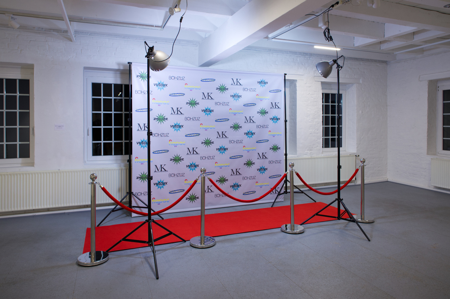 Querformatiges Bild eines weißen Raumes mit dunklen Fenstern und einem grauen Boden. Vor der Fensterwand ist eine Plane mit unterschiedlichen, sich wiederholenden Logos zu sehen. Davor ein roter Läufer, eine Absperrung mit roten Kordeln und zwei Lampen, welche die Plane  mit einem leichten Farbstich beleuchten.