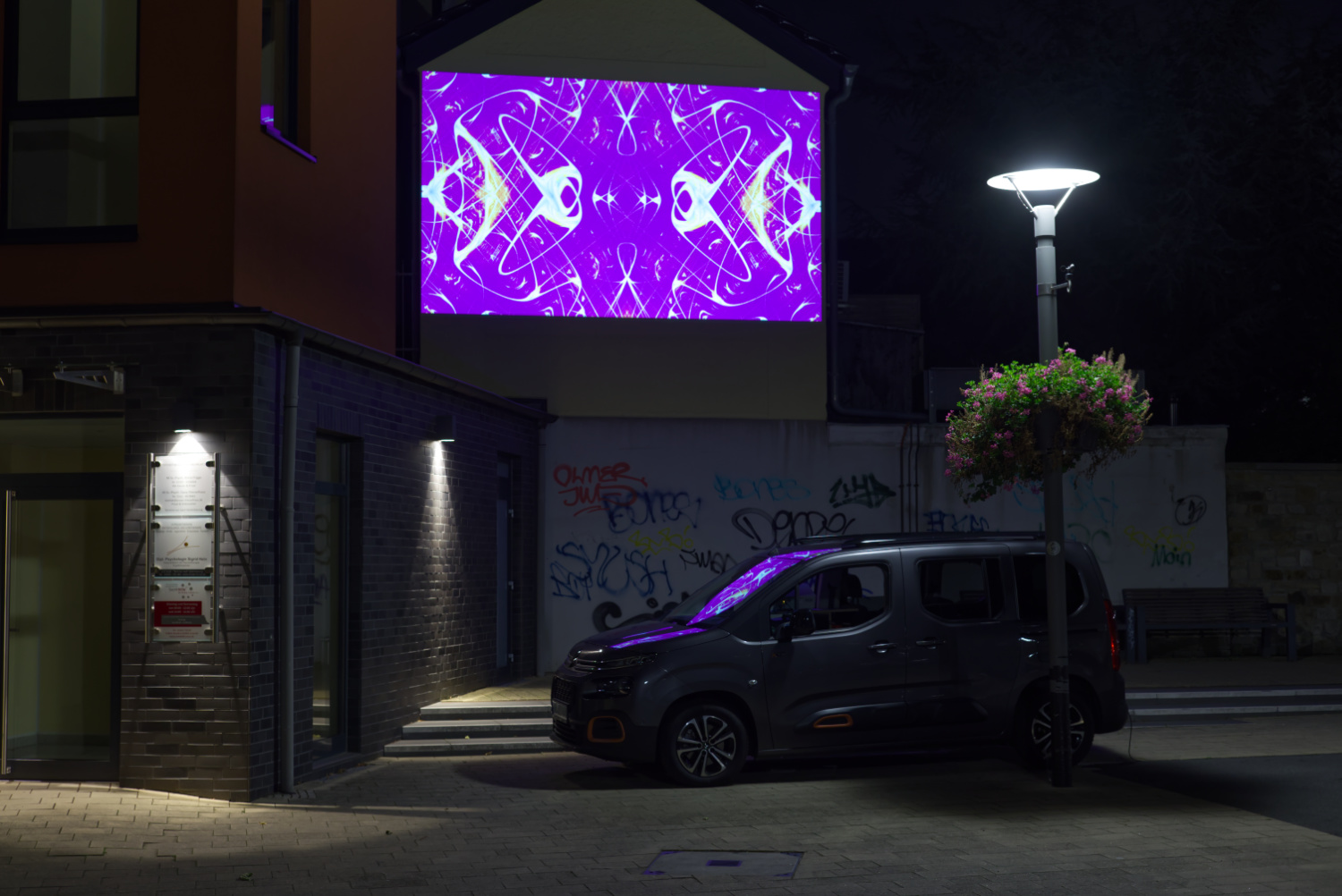Querformatiges Farbfoto. Nachtaufnahme einer Ecke eines Platzes, rechts eine Laterne, drunter steht ein Auto. Auf der Wand eines Gebäudes sieht man seltsame symmetrische Formen vor einem violetten Hintergrund.