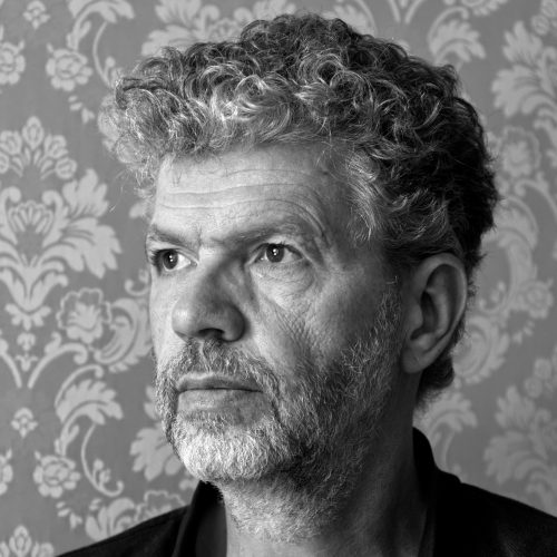 Quadratischen Schwarzweißfoto: ei Porträtaufnahme eines Mannes mit grau melierten Locken und kurzem grau meliertem Bart. Der Blick der dunklen Augen ist gedankenverloren in die Ferne gerichtet.