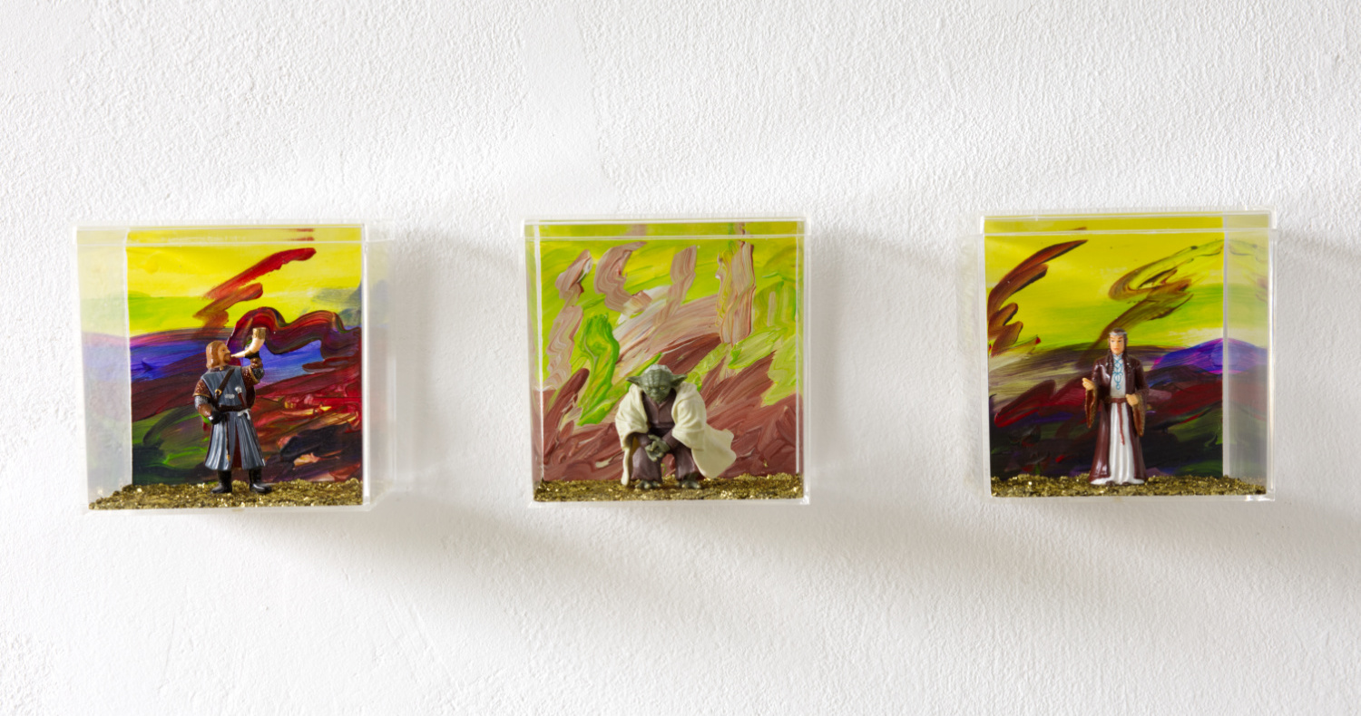 Farbiges Foto im extremen Querformat. An einer weißen Wand hängen drei transparente Kunststoffwürfel nebeneinander. Darin jeweils eine kleine Sammelfigur von Boromir, Yoda und Elrond vor gemalten Hintergründen.  