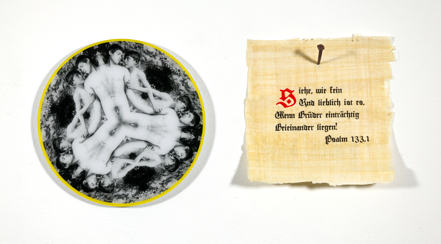 Bild im Querformat. An einer weißen Wand hängt links ein rundes Bild mit gelbem Rand, das zwei nackte Jünglinge als schwarzweiße Fotografie zeigt, wobei das Motiv kaleidoskopartig sechs mal gespiegelt wiederkehrt. Rechts hängt ein quadratisch Stück Papyrus, das mit einem rostigen, geschmiedeten Nagel an der Wand befestigt ist. Darauf steht in Minuskelschrift: "Siehe, wie fein | Und lieblich ist es, | Wenn Brüder einträchtig | Beieinander liegen! | Psalm 133.1"