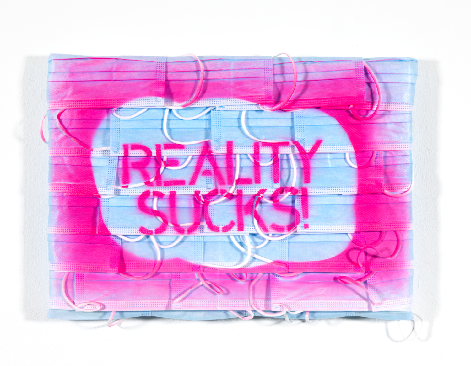 Farbiges Foto im Querformat. An einer weißen Wand hängt ein Bild aus blauen Einwegmasken. Darauf steht in rosa Schrift: "Reality Sucks!" 