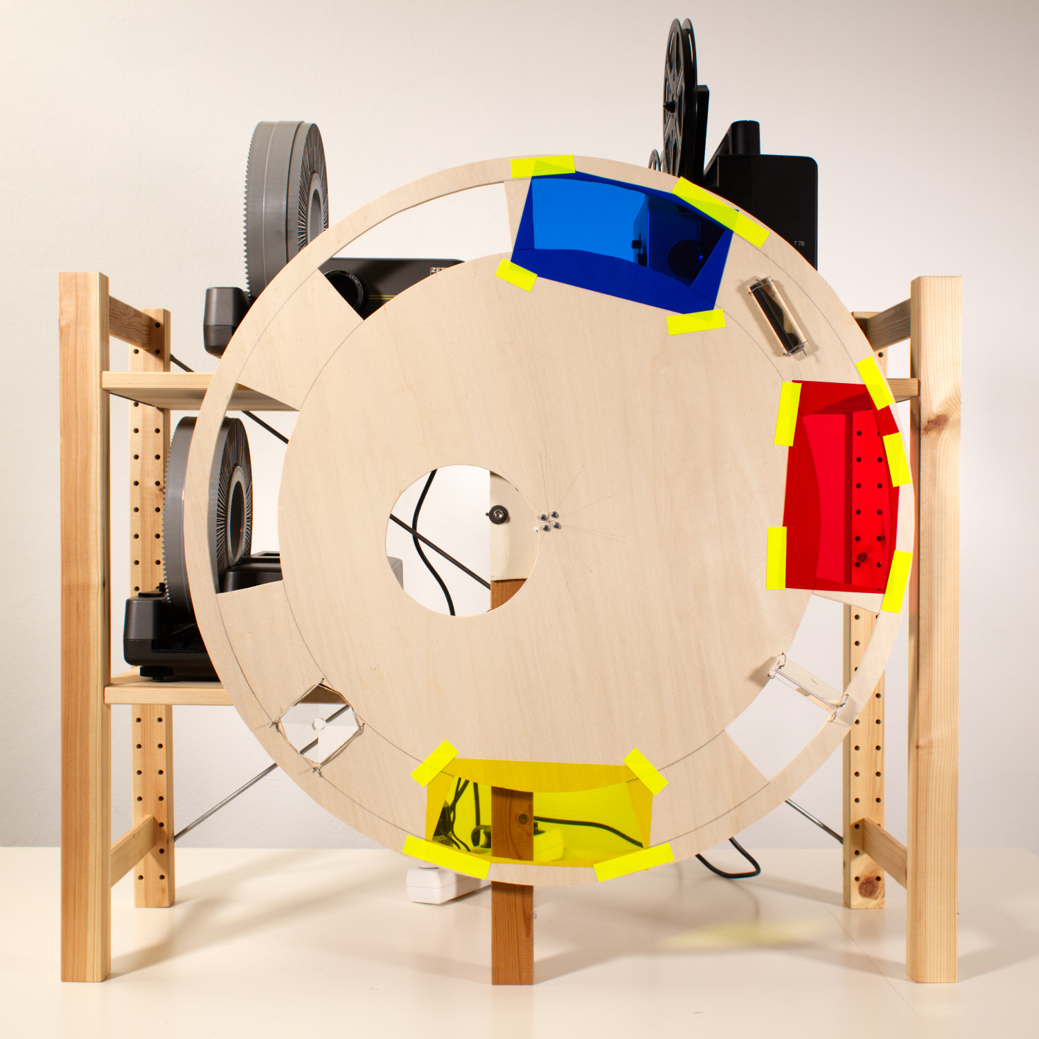 Quadratisches Farbbild eines hölzernen Regals mit zwei Diaprojektoren und einem Super8-Projektor, vor das Regal wurde ein große rund Holzscheibe montiert, darin sind unterschiedliche Löcher, die zum Teil mit farbigen Folien überklebt wurden oder mit Prismen darin.