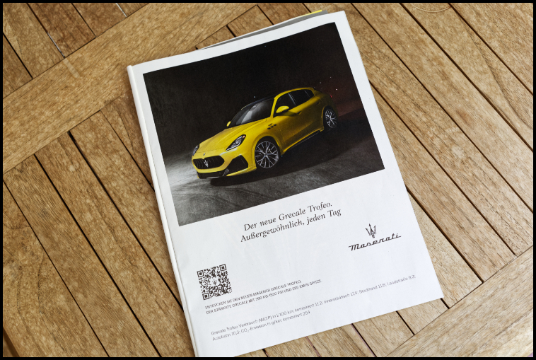 Querformatiges Farbfoto, das ein Magazin auf einem Holztisch zeigt. Aufgeschlagen ist die Ganzseitige Anzeige für einen gelben Maserati SUV. 