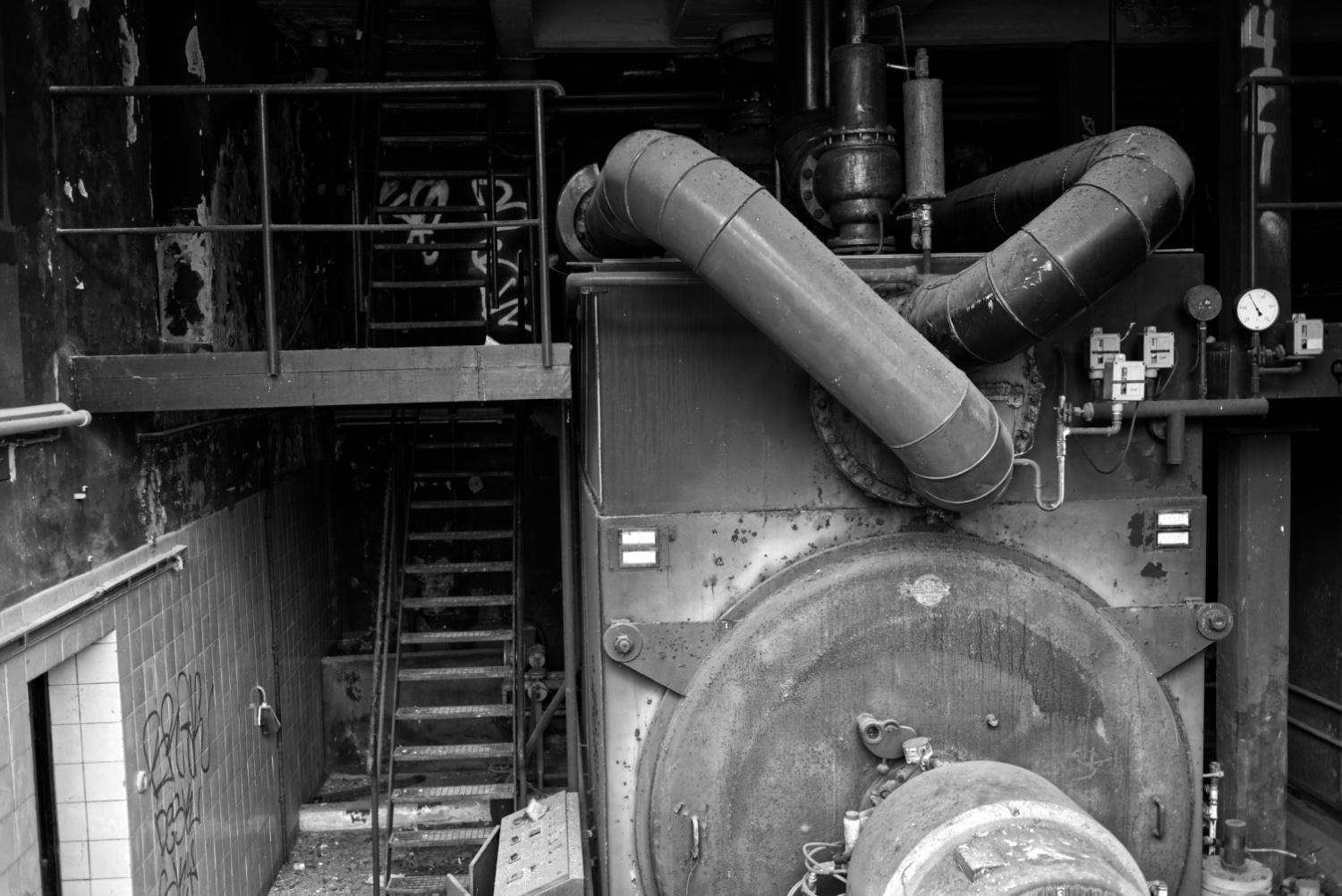 Schwrzweißfoto im Querformat. Die rechte Bildhälfte wird von einer Großen Maschine, vielleicht einem Ofen, dominiert. Links eine Treppe und eine Galerie.