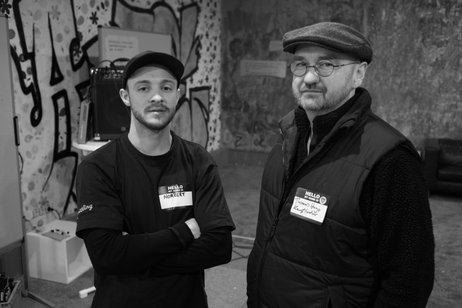 Schwarzweißfoto im Querformat, das zwei Männer mit kurz geschorenen Bärten zeigt, der linke trägt ein Basecap, der Rechte eine Schiebermütze. Beide tragen Namensschilder und zeigen die Andeutung eines Lächelns.