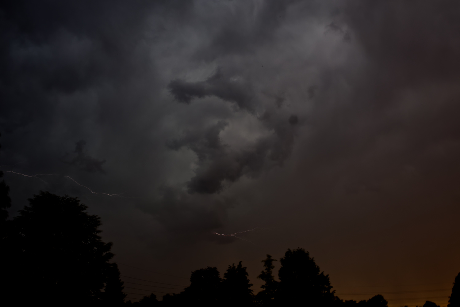 Querformatiges Farbfoto eines dramatischen Abendhimmels mit zwei schwachen Blitzen. Am unteren Bildrand Bäume als Schattenriss.