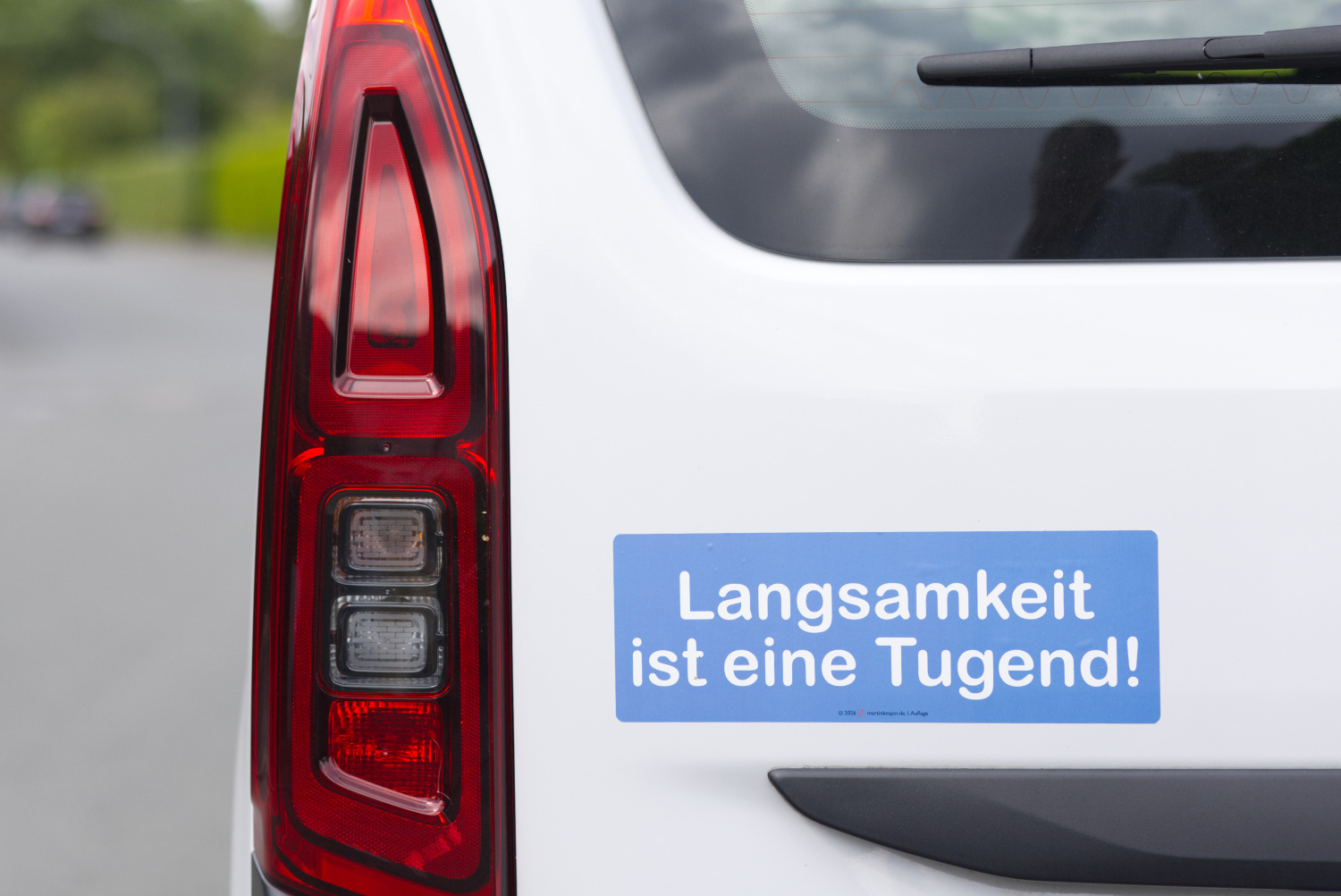 Querformatiges Farbfoto, das zu drei vierteln auf der rechten Seite ein Detail der Rückseite eines weißen Autos zeigt. Neben den linken Rücklichtern, unter der Heckscheibe und über der Kennzeichenbeleuchtung prangt ein blauer, querformatiger Aufkleber, auf dem in weißen, fetten und abgerundeten Zeichen folgender Text steht: „Langsamkeit ist eine Tugend!“